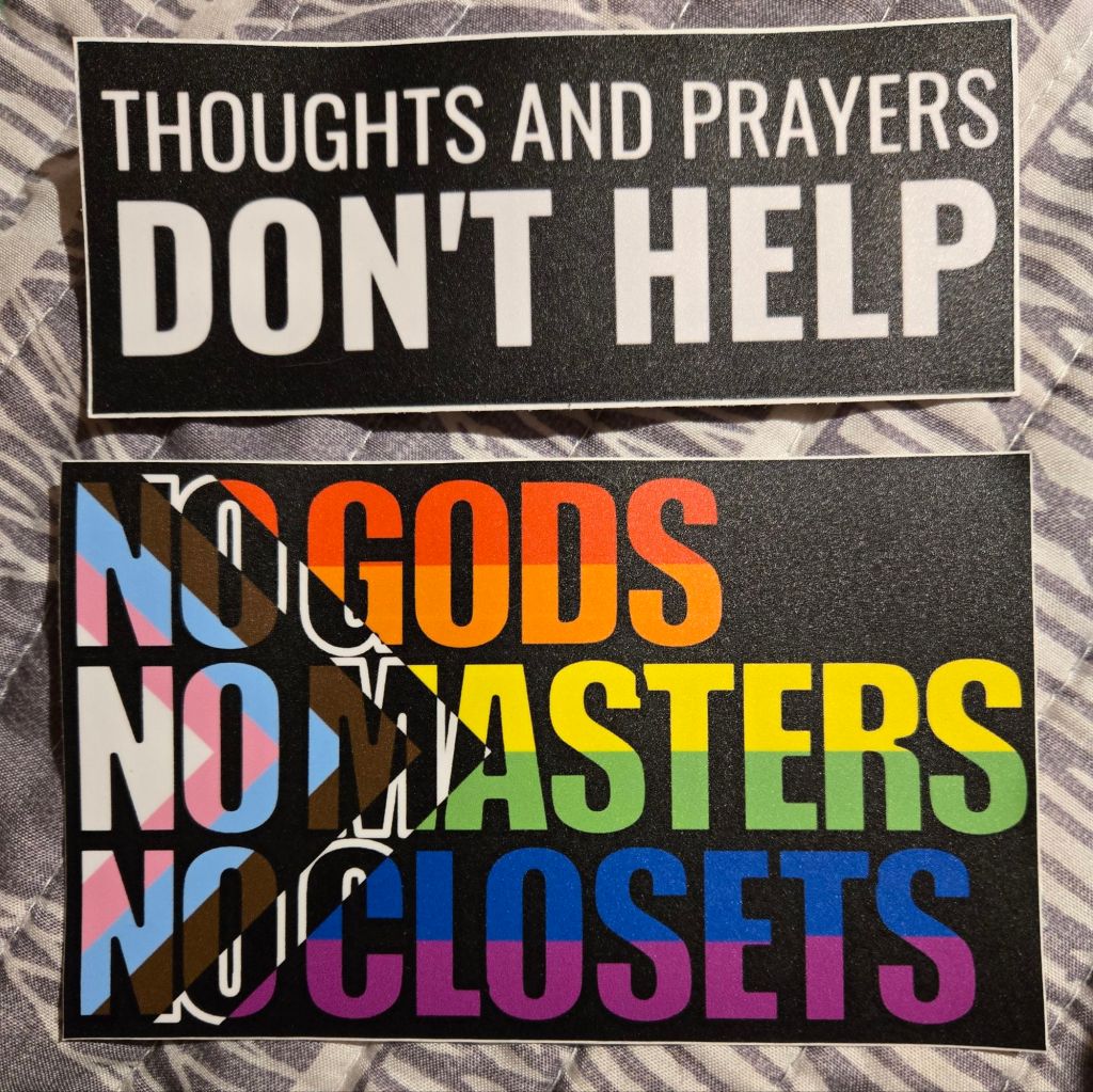 Two large stickers, one in black and white saying "THOUGHTS AND PRAYERS DON'T HELP" and another in the Progress Pride flag colors saying "NO GODS NO MASTERS NO CLOSETS"