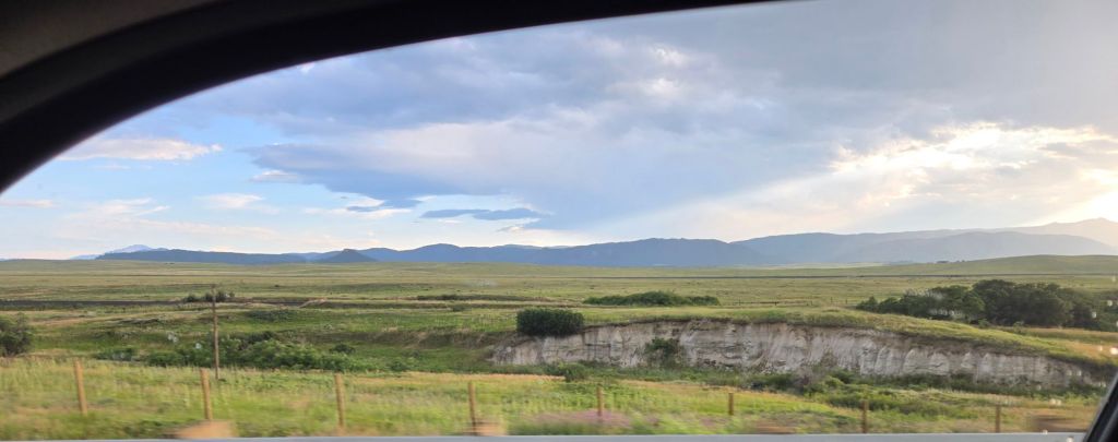 A large, grassy plain with hazy mountains in the distance and a bright blue sky brushed with clouds, tinged yellow with the sunset on the right side.