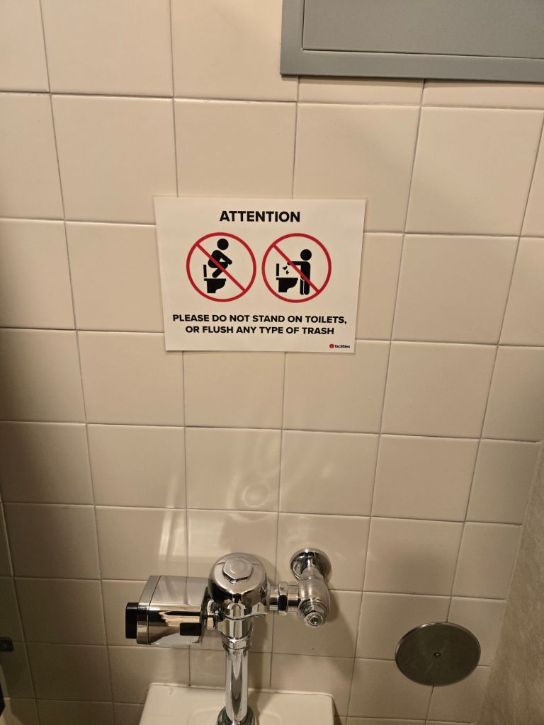 A toilet stall with a sign on the wall saying "ATTENTION: please do not stand on toilets, or flush any type of trash." Above the text are two crossed-out pictograms demonstrating the forbidden acts.