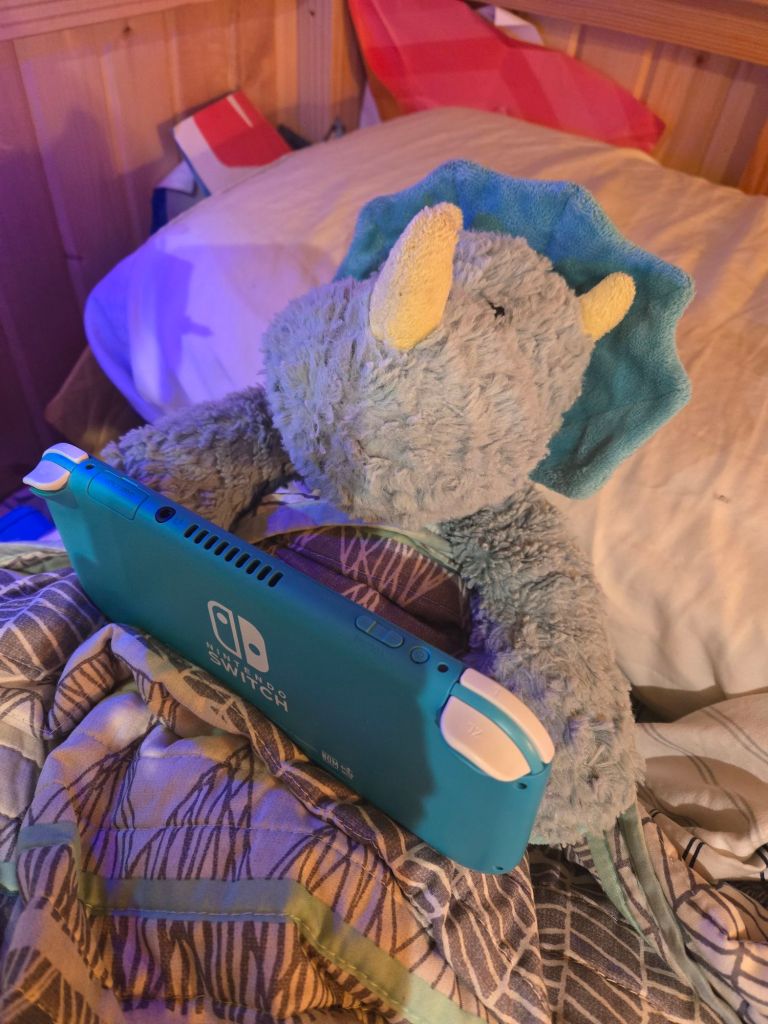 A stuffed triceratops propped against a pillow in an adult bed, tucked in and holding a Nintendo Switch Lite.