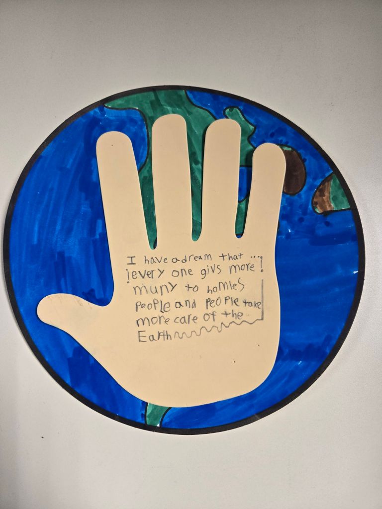 A paper cutout of a child's hand, glued onto a circle of paper colored to look like the Earth. Written on the hand is a child's dream for people to take better care of the homeless and the planet.